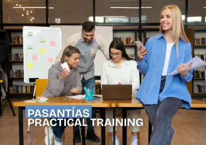 Chica sonriendo mientras sostiene su celular, con tres personas al fondo trabajando en laptops, representando un ambiente de pasantías y formación práctica.